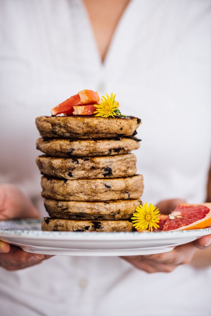 Woman holding a stack of egg free pancakes with chocolate chips on a plate with flowers and grapefruit wedges.