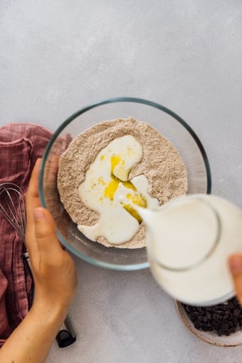 Woman pouring milk into the dry ingredients mixture in a glass bowl to make fluffy egg free pancakes.