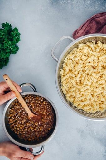 Woman holding cooked green lentil bolognese sauce. Cooked pasta in a strainer on the side.