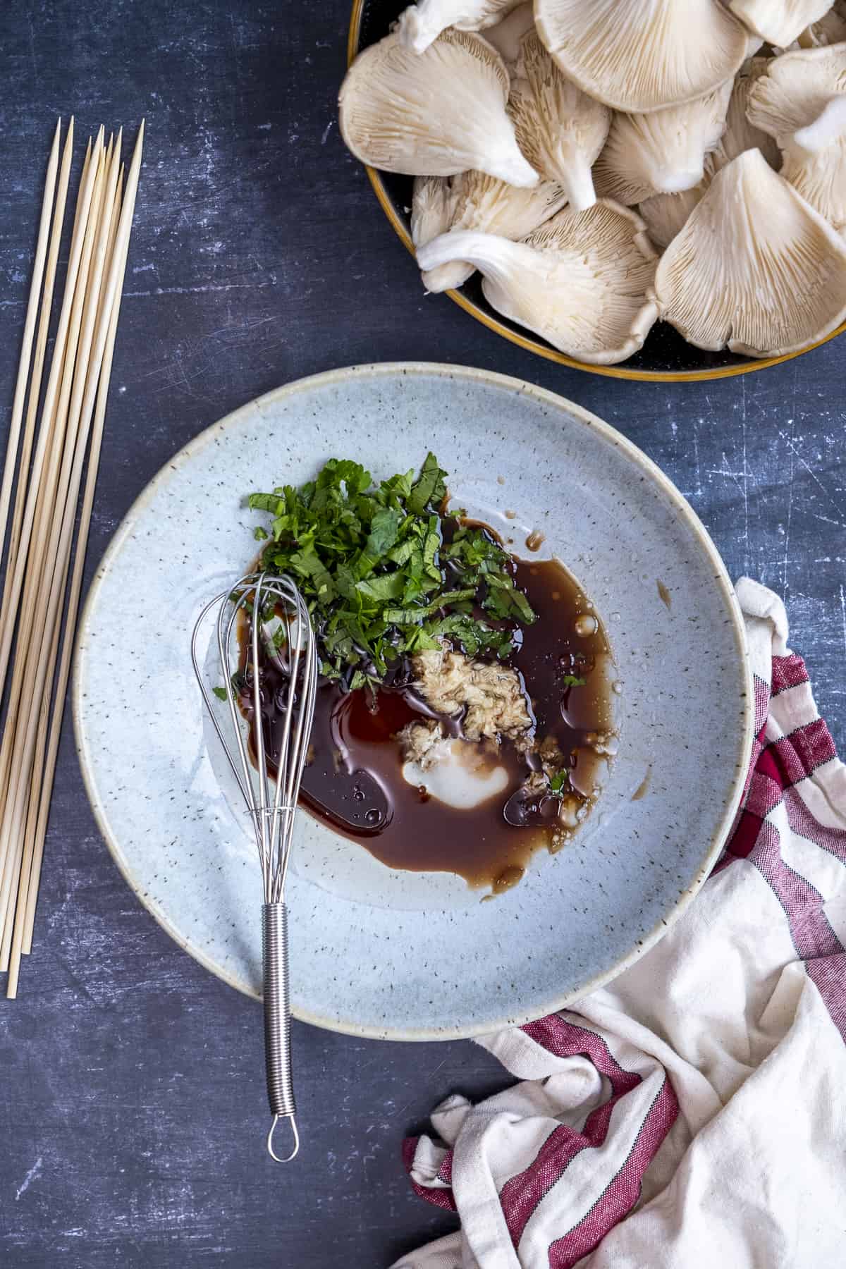 Marinade ingredients in a blueish bowl, a hand whisk inside it, wooden skewers and oyster mushrooms on the side.