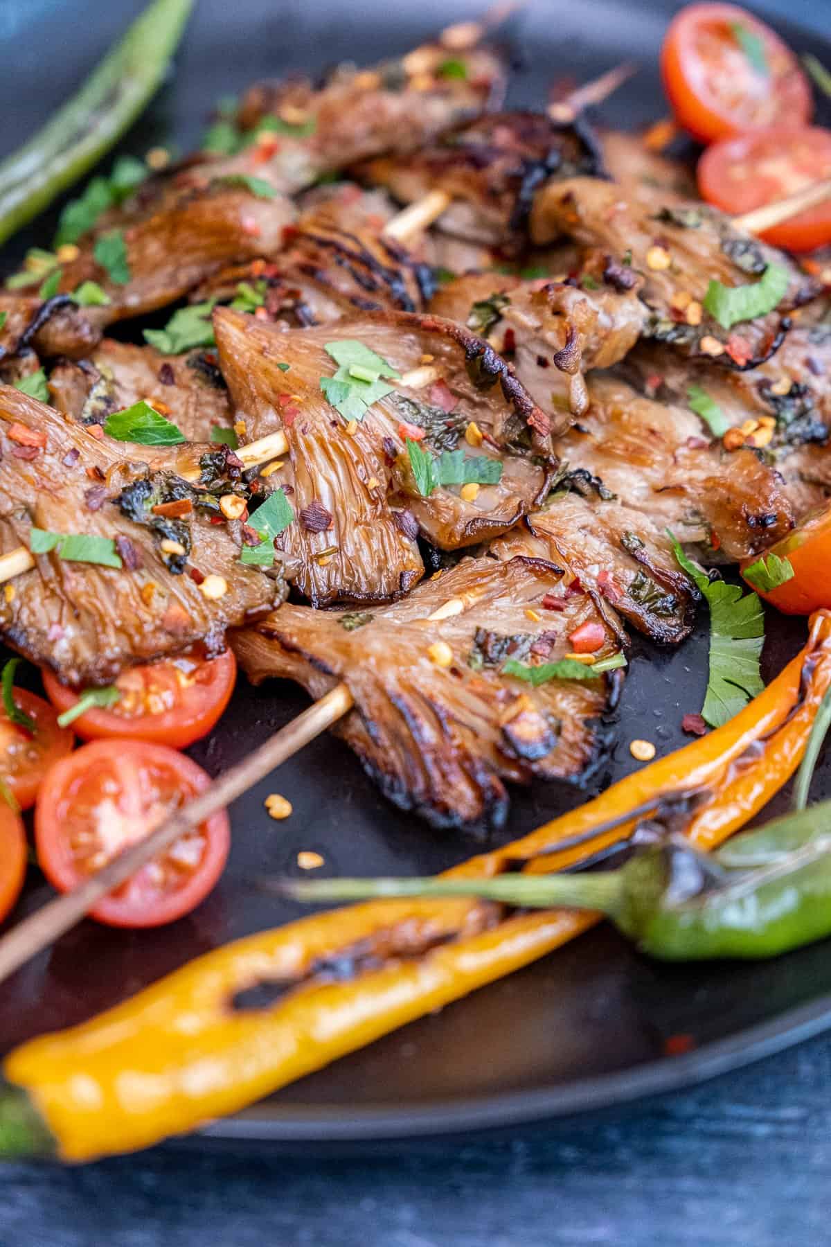 Grilled oyster mushrooms on skewers served with grilled peppers and tomatoes on a black plate.