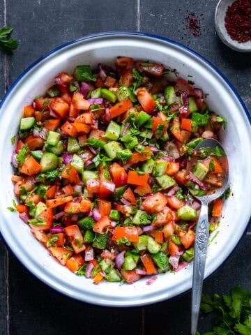 A bowl of Çoban Salata with diced tomatoes, cucumbers, peppers, onions, and parsley, tossed in olive oil, lemon juice, and sumac. A spoon rests inside, with sumac and parsley nearby.