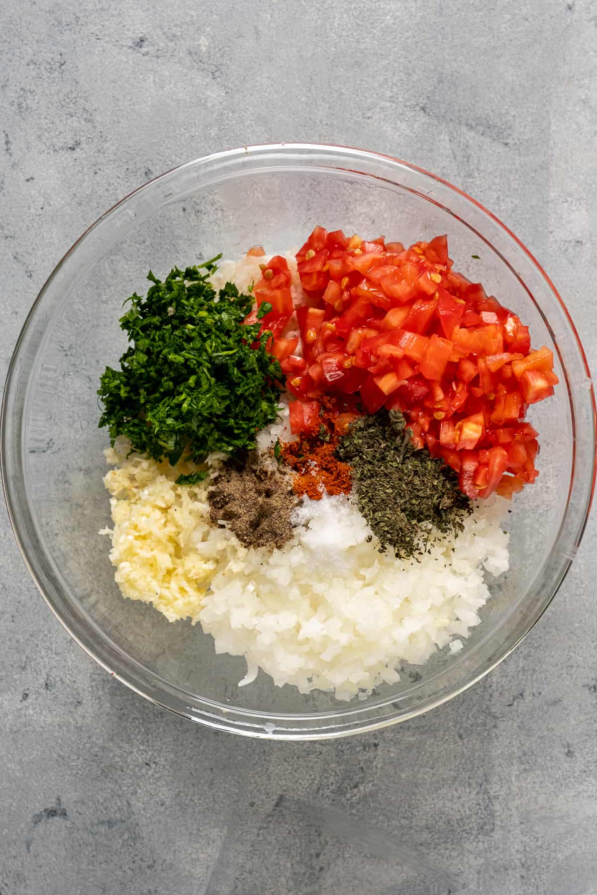 Rice, chopped garlic, onion, tomatoes, parsley and spices in a glass mixing bowl.