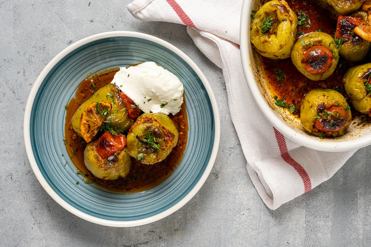 Stuffed green peppers served on a bluish plate with its tomato sauce and some yogurt on the side. The baking pan with the remaining dish accompanies.