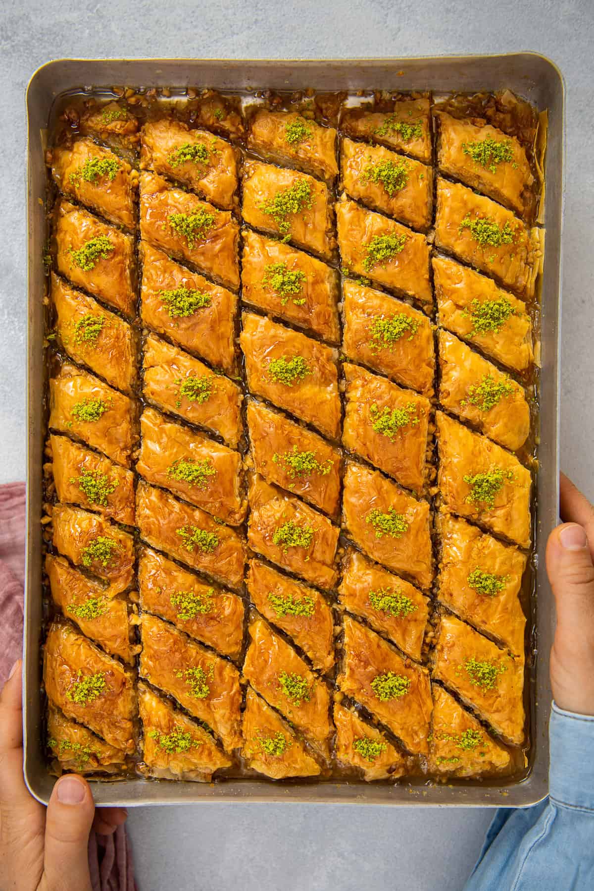 Freshly baked Turkish baklava in a tray with syrup and pistachio topping, held in hands.
