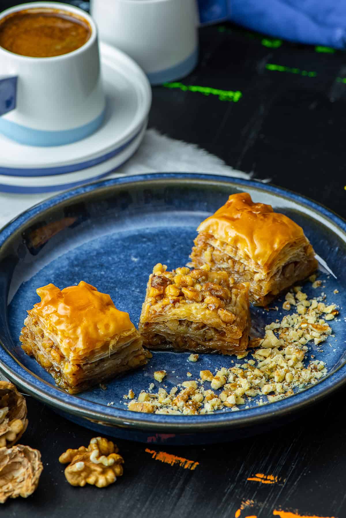 Slices of Turkish walnut baklava on a blue plate, topped with syrup and crushed walnuts, served with Turkish coffee in the background.