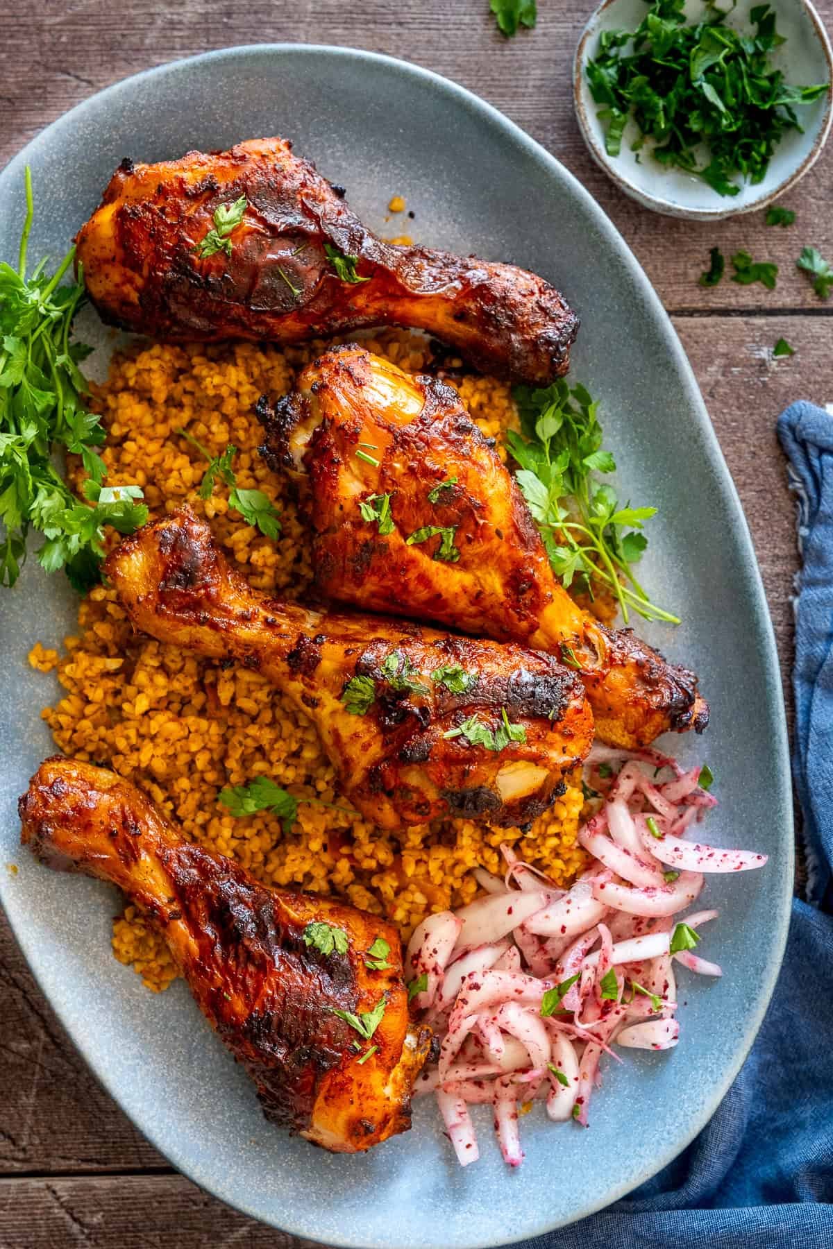 Close-up of Turkish chicken legs served on bulgur pilaf with sumac onions and fresh parsley on a blue platter.