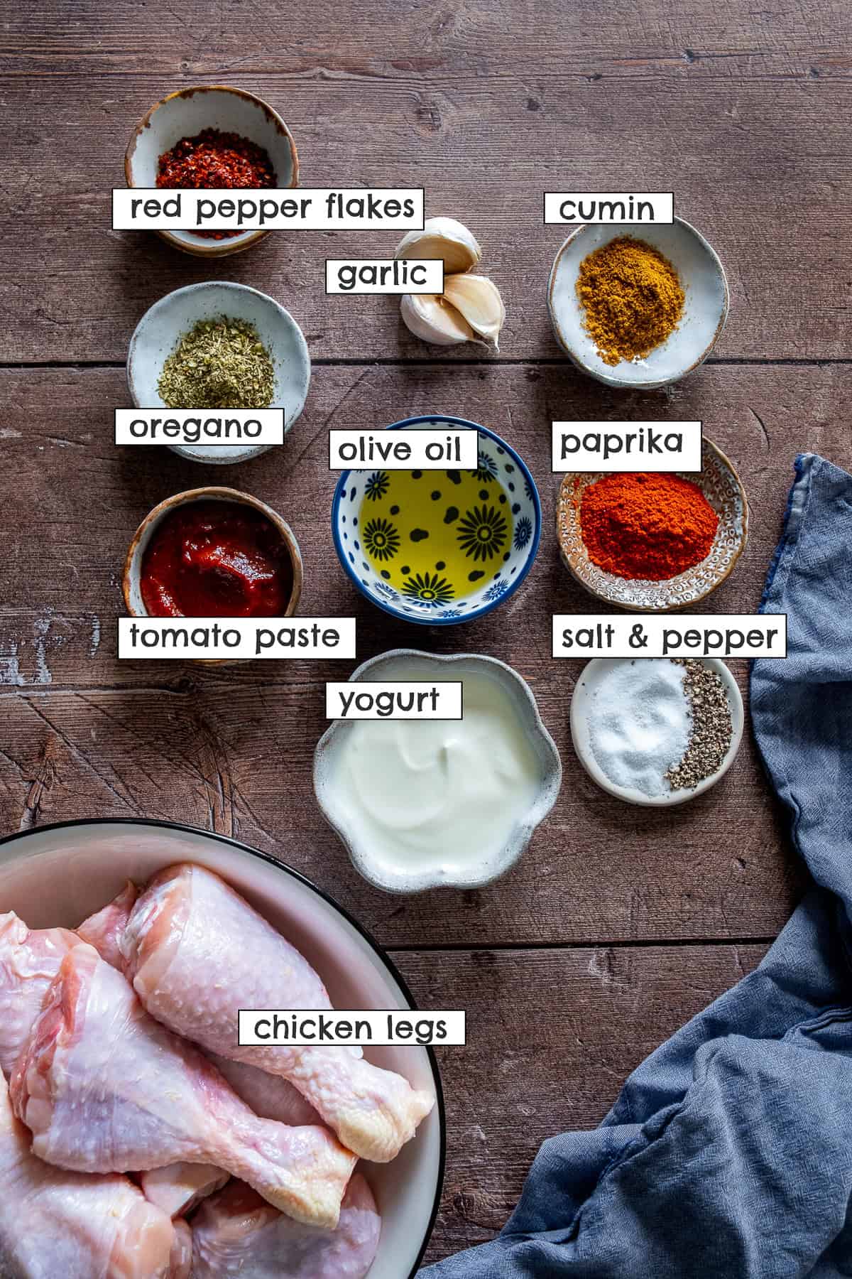 Ingredients for Turkish chicken legs laid out on a wooden surface, including chicken drumsticks, yogurt, tomato paste, olive oil, garlic, and spices like paprika, cumin, oregano, and red pepper flakes.