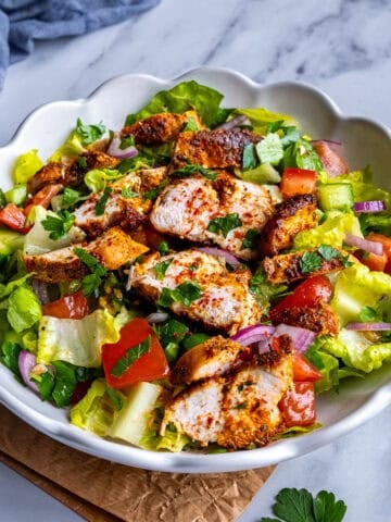 Lettuce salad with tomatoes, cucumbers, onions and parsley topped with cooked chicken breast slices in a white bowl.