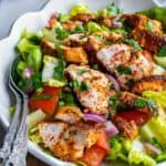 Grilled Turkish spiced chicken salad served in a white bowl with lettuce, tomatoes, red onion, and fresh herbs, topped with sliced chicken and red pepper flakes.