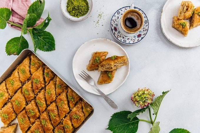 Turkish coffee in a traditional Turkish coffee cup, two slices of turkish baklava on a ceramic plate, a baking sheet full of newly made baklava 