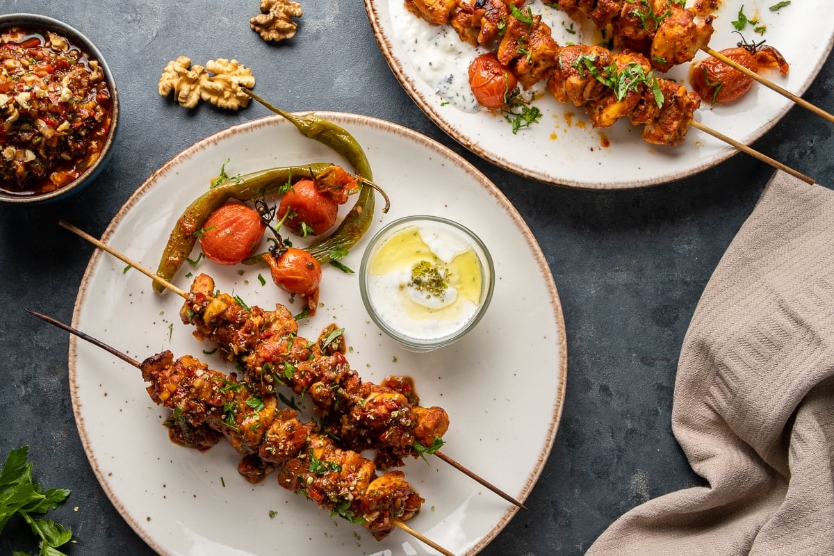 Chicken skewers served on two white plates, roasted tomatoes and peppers and a small bowl of yogurt sauce on the same plates.