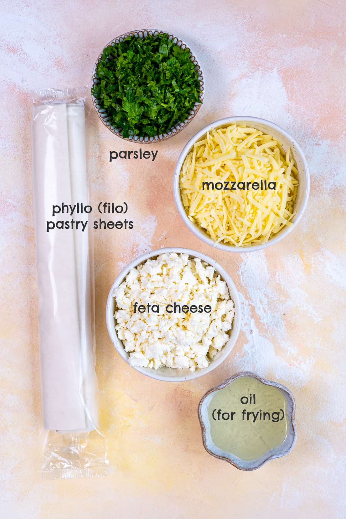 Phyllo dough, grated mozzarella, crumbled feta cheese, parsley and oil in separate bowls on a light background.