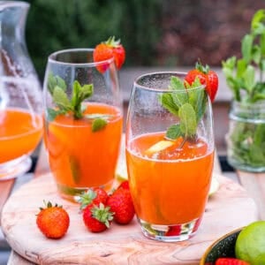 Strawberry lemonade in two tall glasses garnished with mint leaves and strawberries on a wooden board with an outside background.