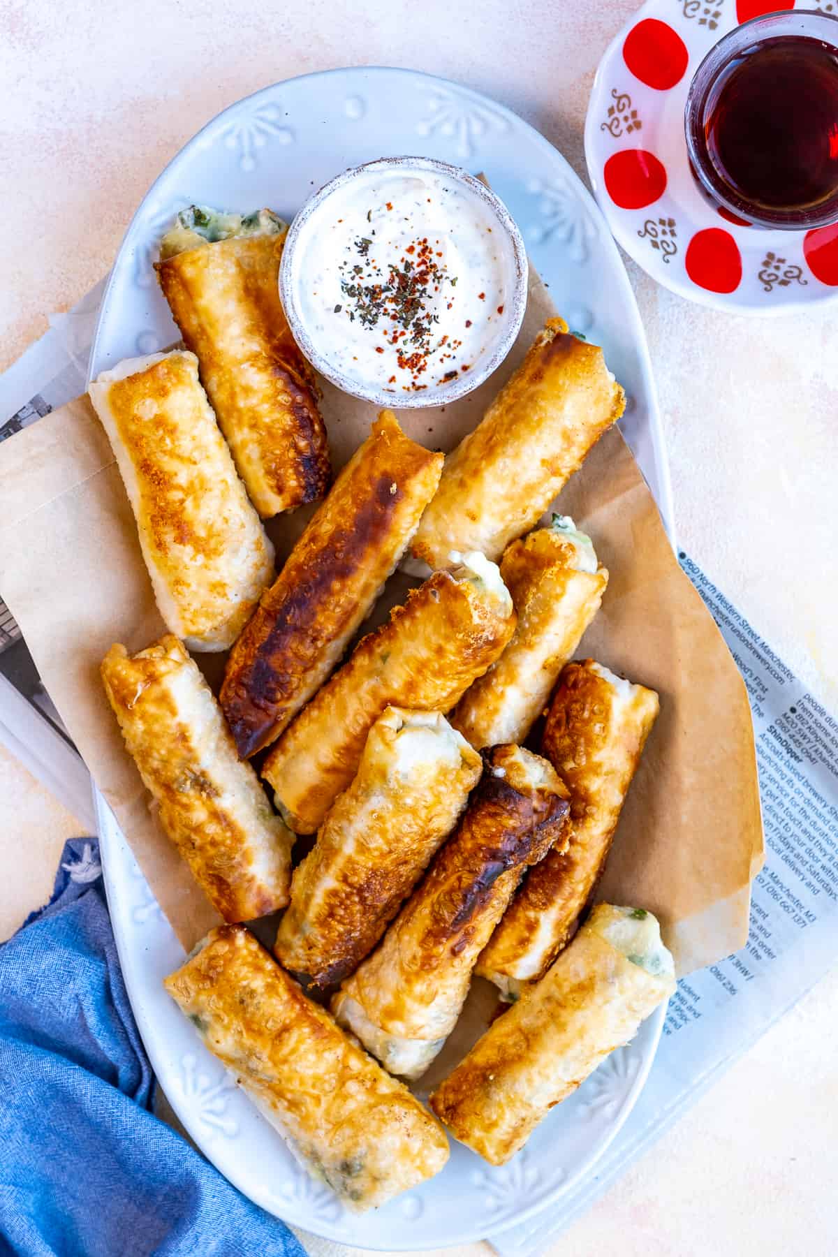 Cheese rolls on a white plate served with a yogurt sauce in a small bowl on the side.