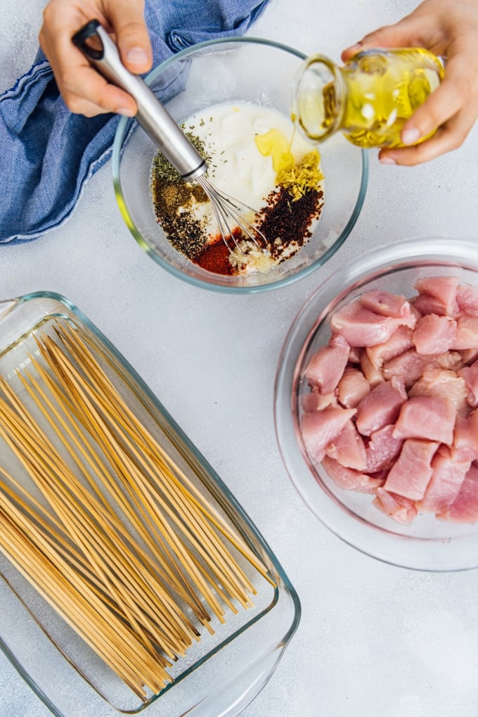 Chicken cubes in a bowl, hands preparing the marinade in another bowl and wooden skewers soaked in water in a pan.