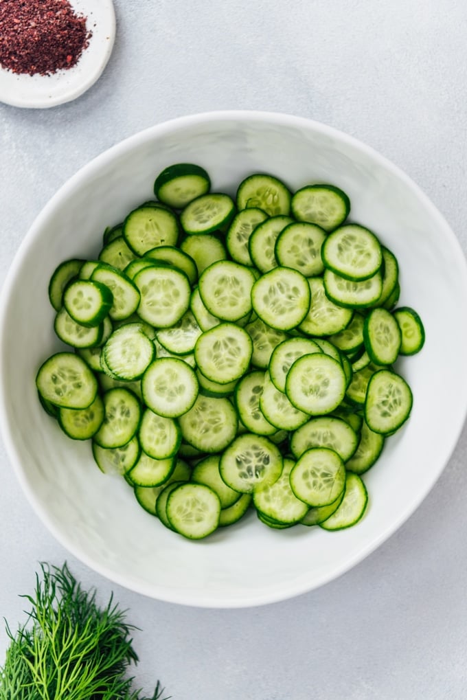 The BEST Creamy Cucumber Salad with onions and yogurt dressing. No mayonnaise. This will be your new summer hit! #creamycucumbersalad #cucumbersalad #saladwithoutmayo #creamycucumbers Sliced cucumbers in a white salad bowl for creamy cucumbers recipe
