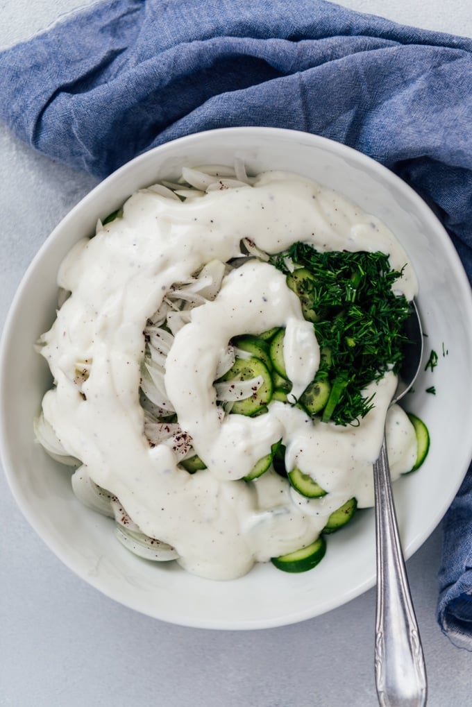 The BEST Creamy Cucumber Salad with onions and yogurt dressing. No mayonnaise. This will be your new summer hit! #creamycucumbersalad #cucumbersalad #saladwithoutmayo #creamycucumbers Creamy cucumbers and onions with dill and Greek yogurt in a white bowl