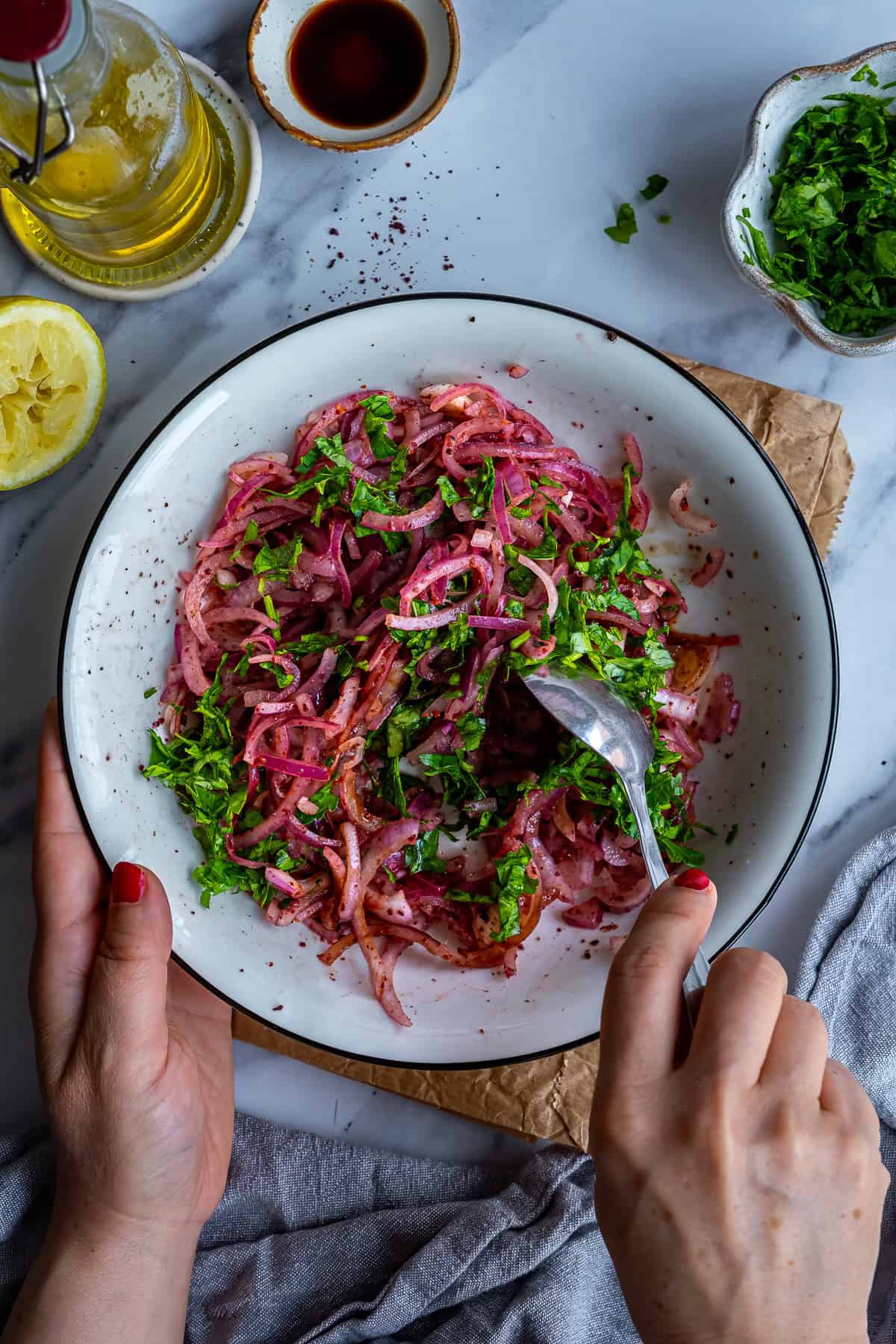 Hands mixing Turkish sumac onions with parsley, lemon juice and pomegranate molasses in a white bowl.