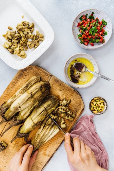 Hands chopping roasted whole eggplants on a wooden board, salad dressing and chopped tomatoes on the side.