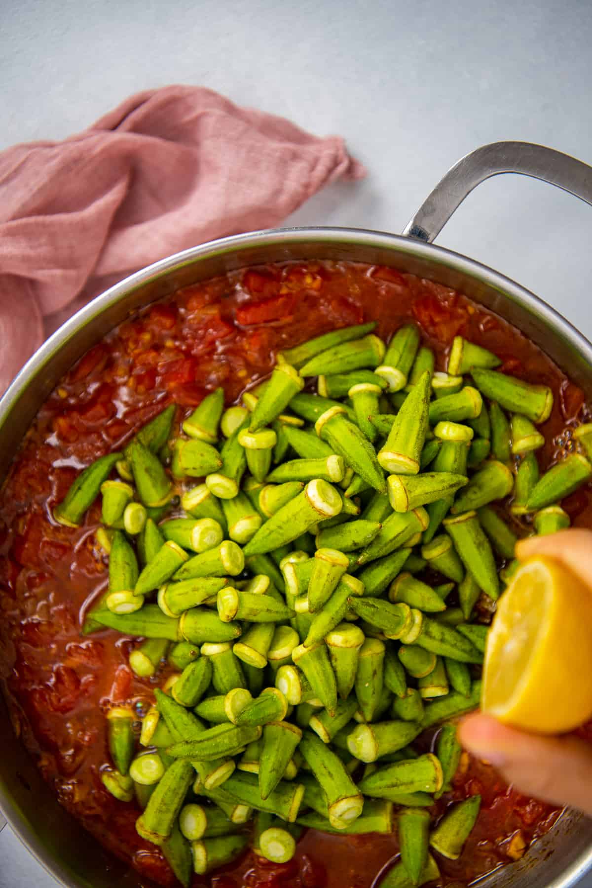 Fresh okra being cooked in a tomato sauce in a pan and hand squeezing lemon over it.