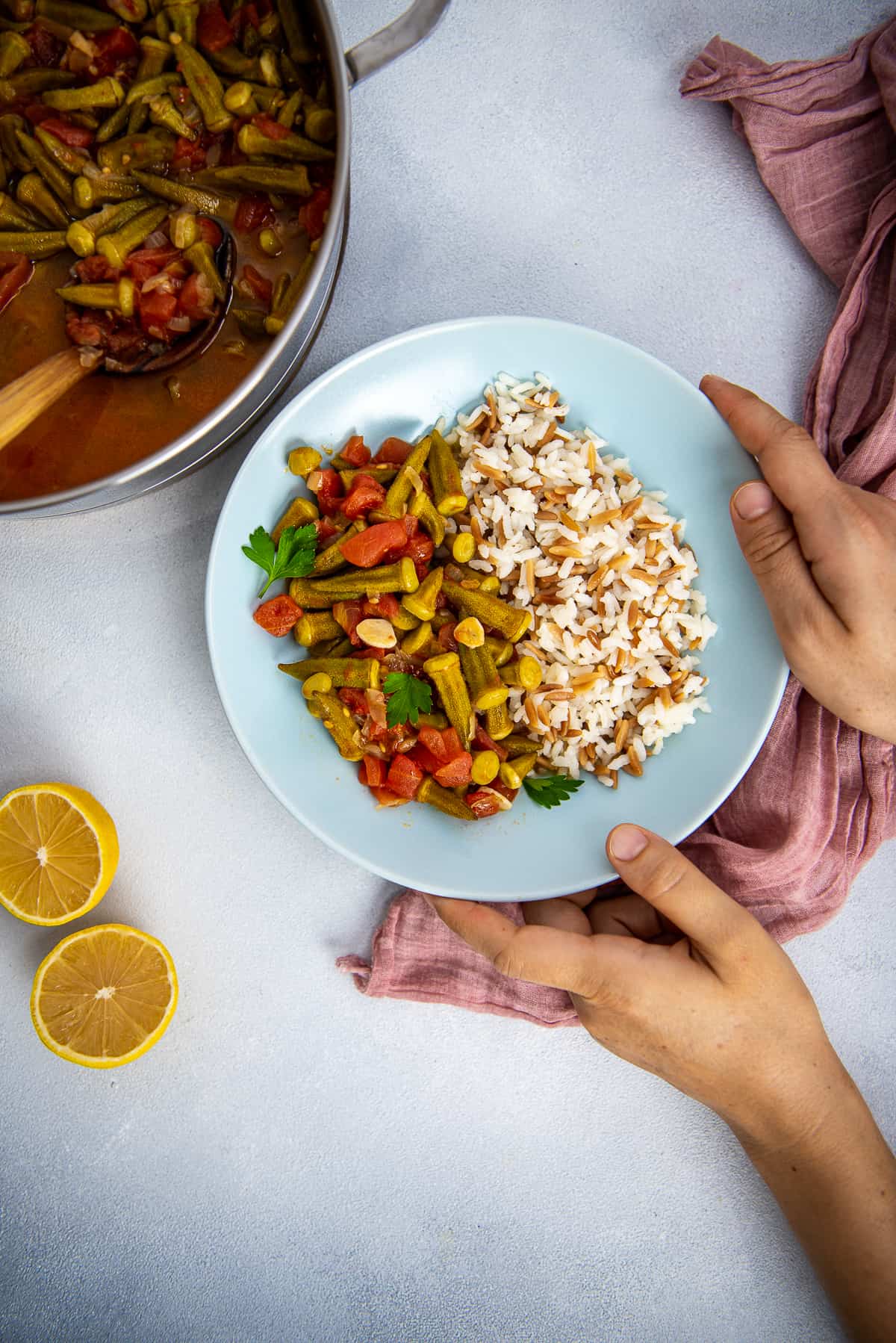 Hands serving bamya paired with rice in a bowl, lemon halves and a pan full of bamya on the side.