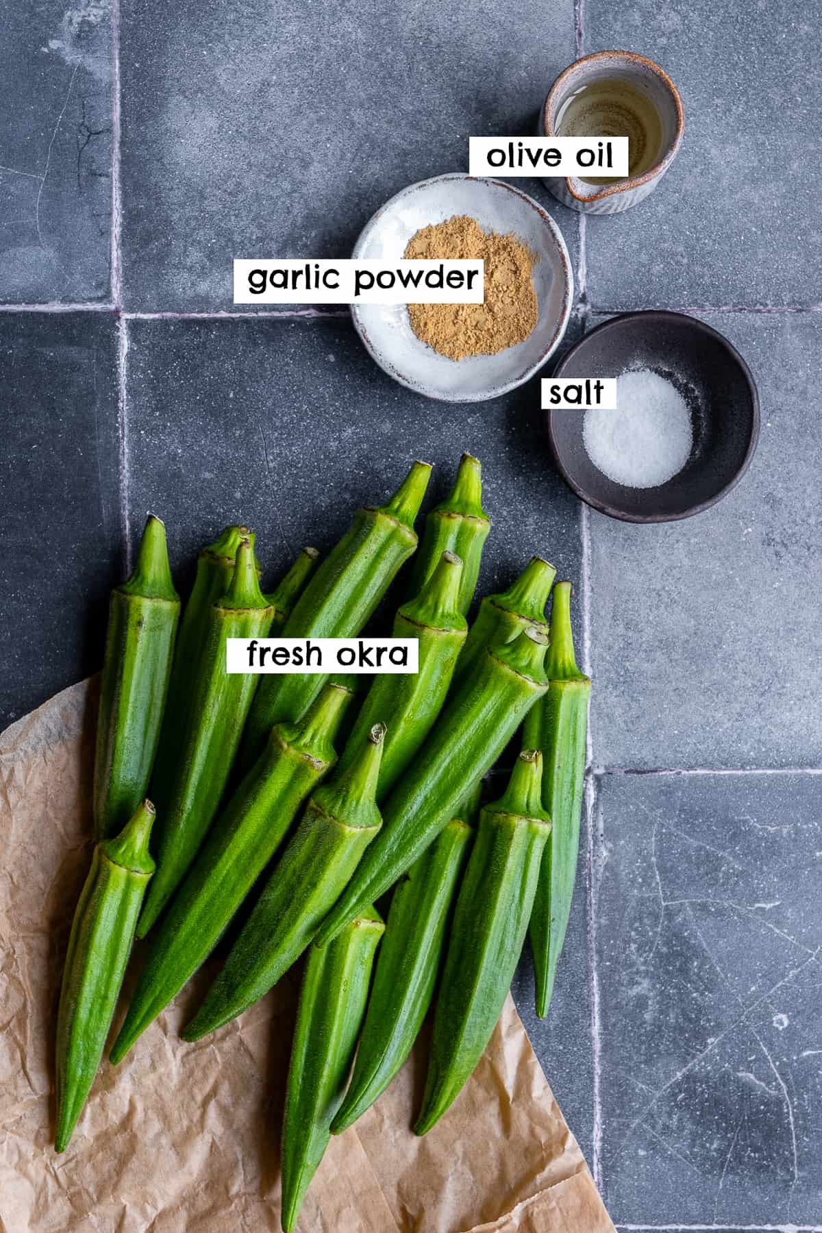 Fresh okra pods, salt, garlic powder and oil on grey tiles.