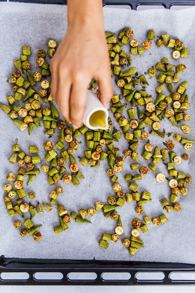 A hand drizzling olive oil over chopped and spiced okras on a baking sheet.