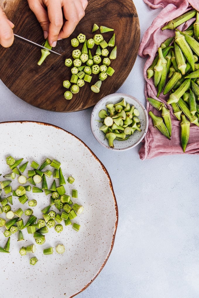 Hands cleaning and chopping okras on a round wooden board, whole okras on the side.