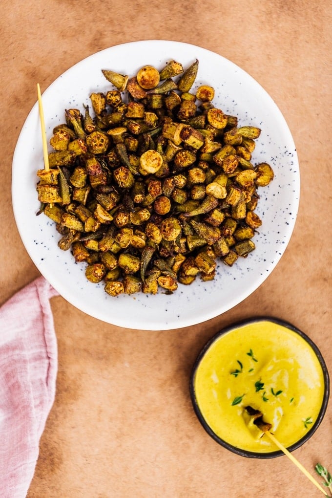 Oven roasted Okra Recipe is for those who hate okra. These crispy roasted okra chips will become their new favorite snack. Healthy and incredibly tasty! #roastedokra #okrarecipe #healthysnack Crispy roasted okra chips in a white ceramic bowl and turmeric tahini dip in a small bowl on the side.