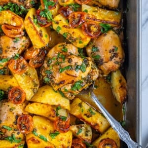Baked chicken thighs, potatoes and cherry tomatoes garnished with parsley in a baking pan and a spoon inside it.
