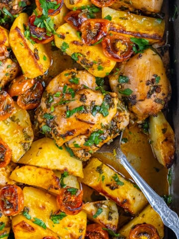 Baked chicken thighs, potatoes and cherry tomatoes garnished with parsley in a baking pan and a spoon inside it.