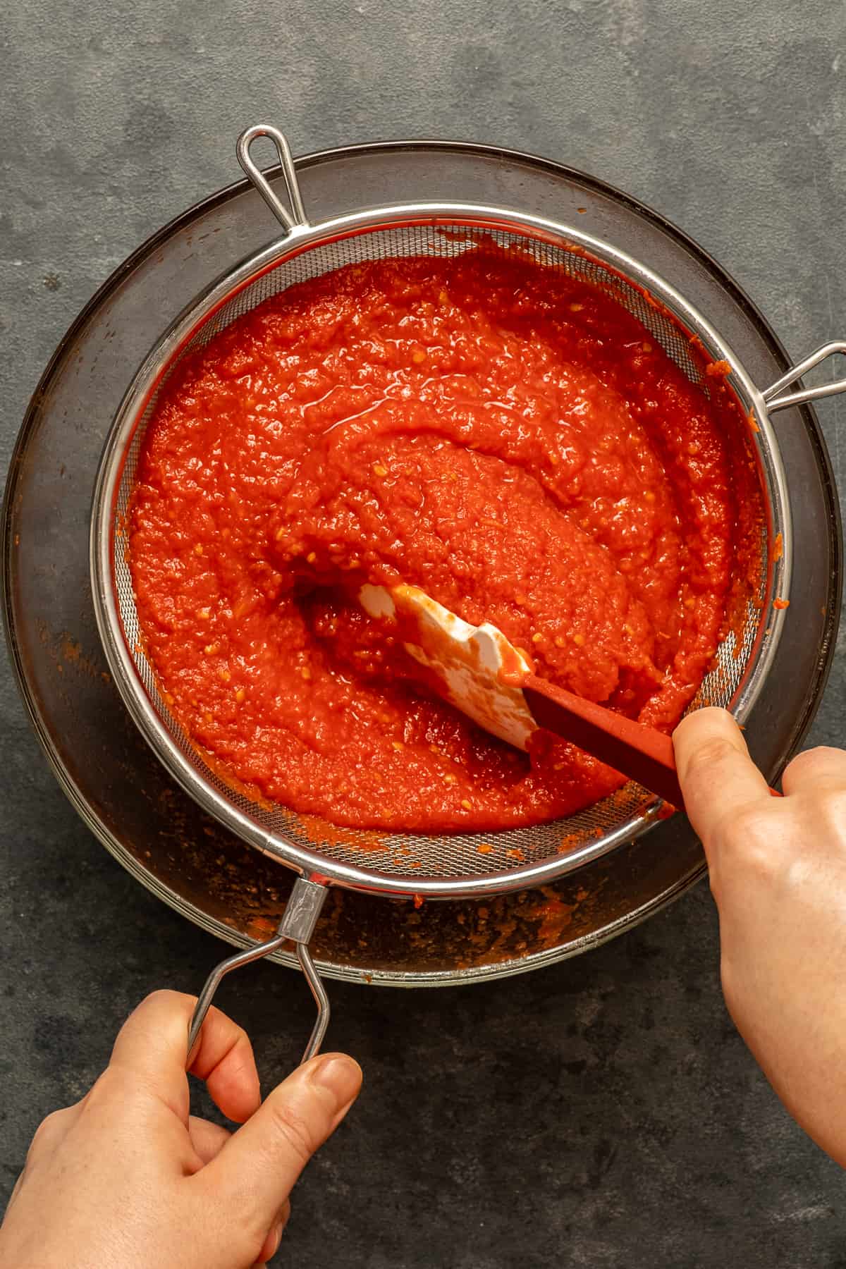 Hands straining cooked tomato puree in a fine mesh strainer.