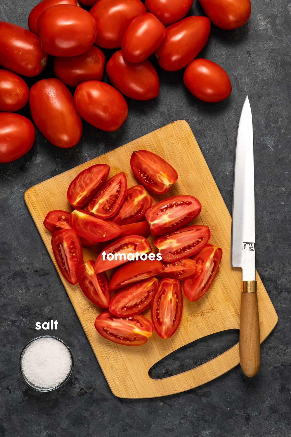 Quarted tomatoes on a wooden cutting board, a knife on it, kosher salt in a small bowl and whole tomatoes on the side.