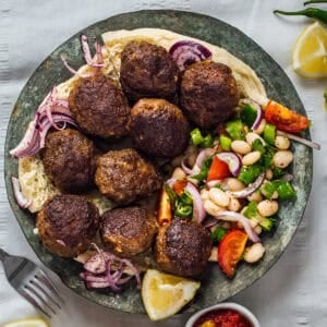 Plate of Turkish kofte (meatballs) served on flatbread with piyaz, sumac onions and lemon wedges.