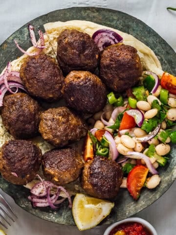 Plate of Turkish kofte (meatballs) served on flatbread with piyaz, sumac onions and lemon wedges.