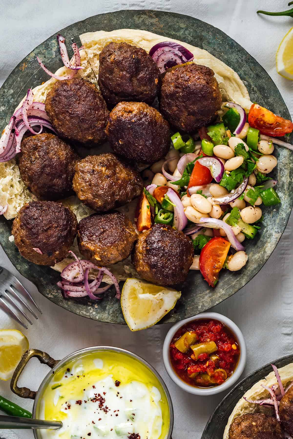 Plate of Turkish kofte (meatballs) served on flatbread with piyaz (Turkish white bean salad), sumac onions, cacık, pickles and lemon wedges.