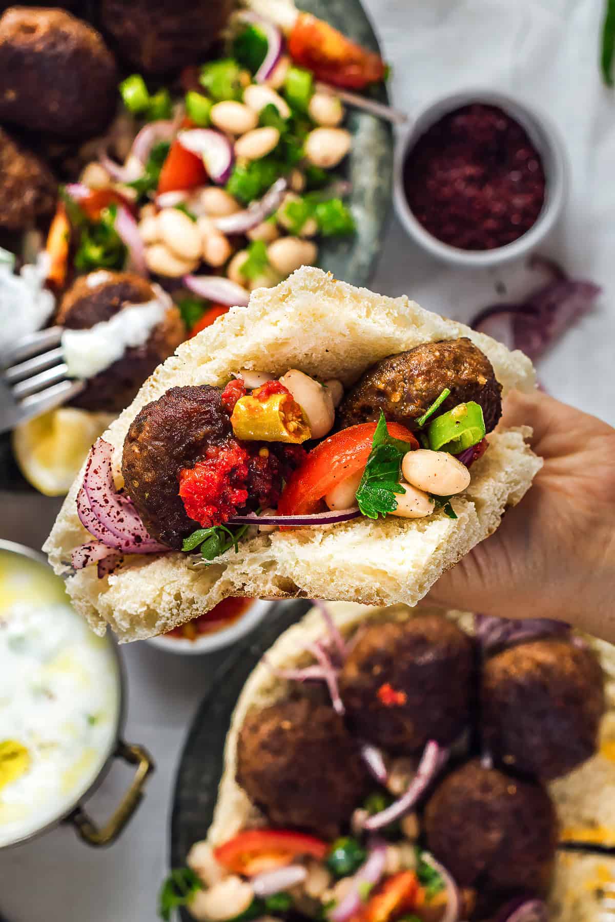 Hand holding a Turkish kofte sandwich in a piece of flatbread filled with kofte, piyaz (Turkish white bean salad), sumac onions, tomatoes and pickles, with Turkish side dishes in the background.