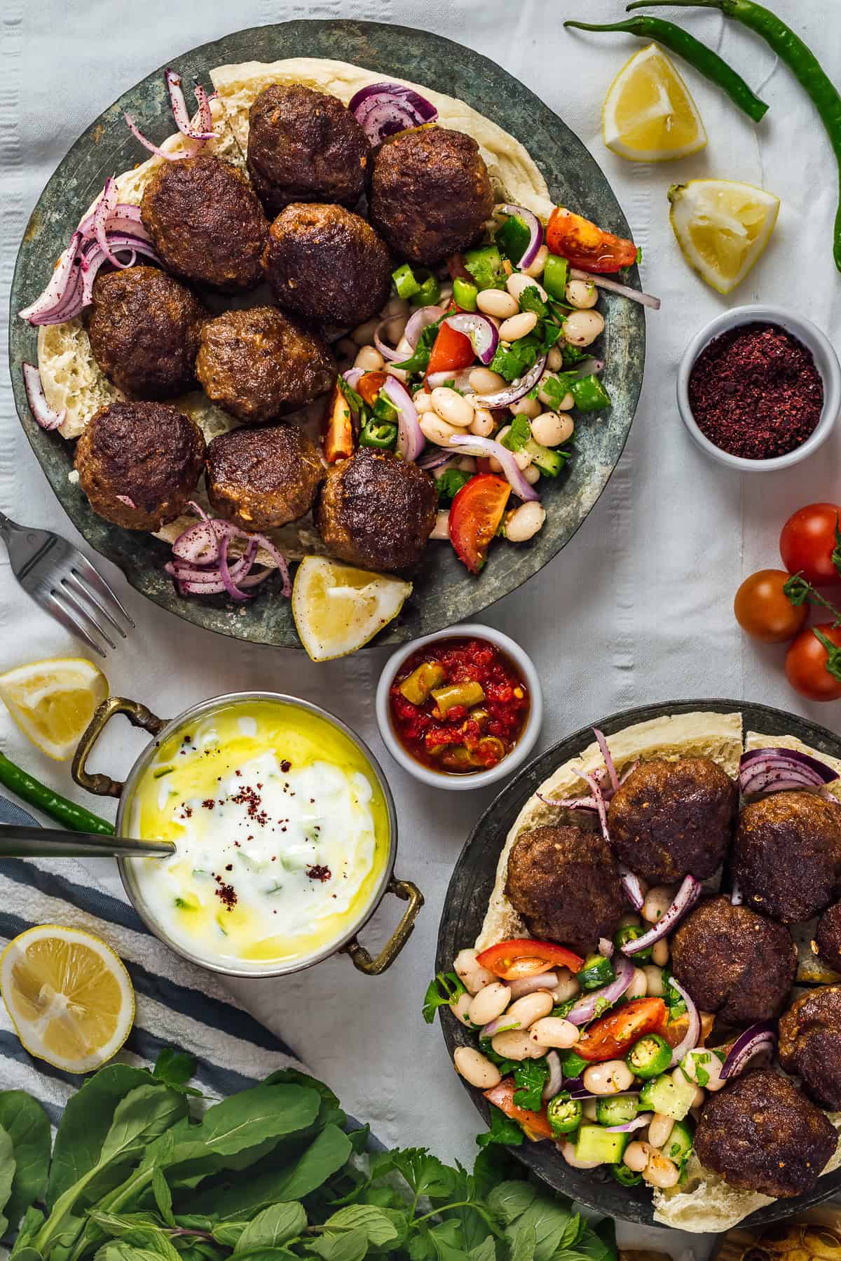 Two plates of Turkish kofte (meatballs) served on flatbread with piyaz (Turkish white bean salad), sumac onions, cacık, pickles and lemon wedges.