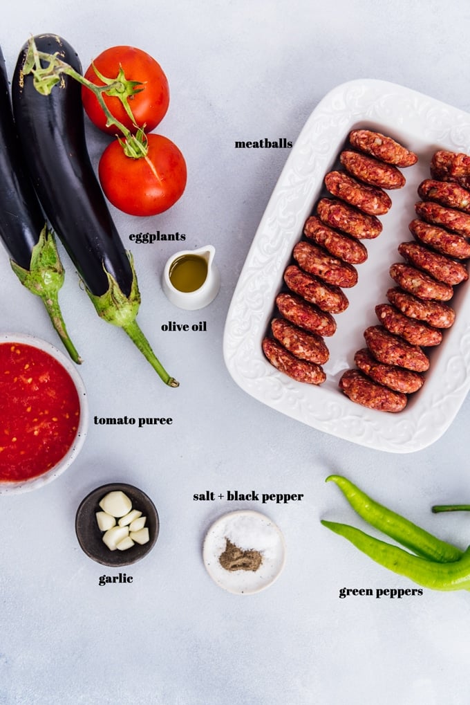 Eggplants, tomatoes, tomato puree, garlic cloves, salt and pepper, green peppers on a grey background and meatballs in a white rectangular pan on the side.