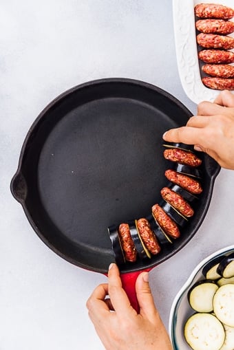 Hands placing eggplant and meatballs in a cast iron skillet