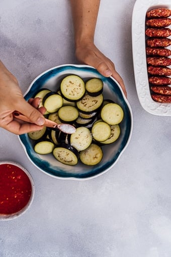 Hands sprinkling salt on sliced eggplants in a ceramic bowl and homemade meatballs on the side.