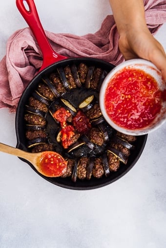 Hands pouring pureed tomato on baked eggplant slices and meatballs in a cast iron skillet.