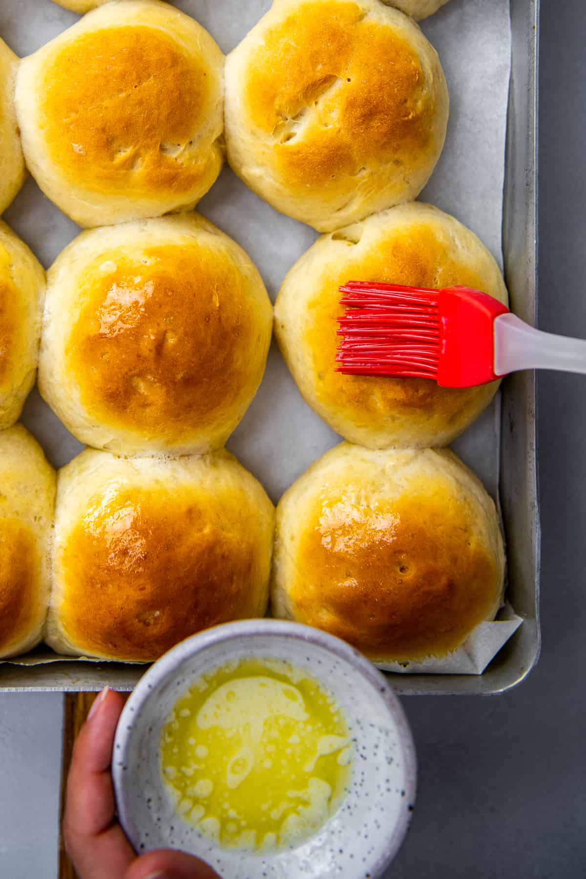 Homemade dinner rolls in a baking tray being brushed with melted butter.