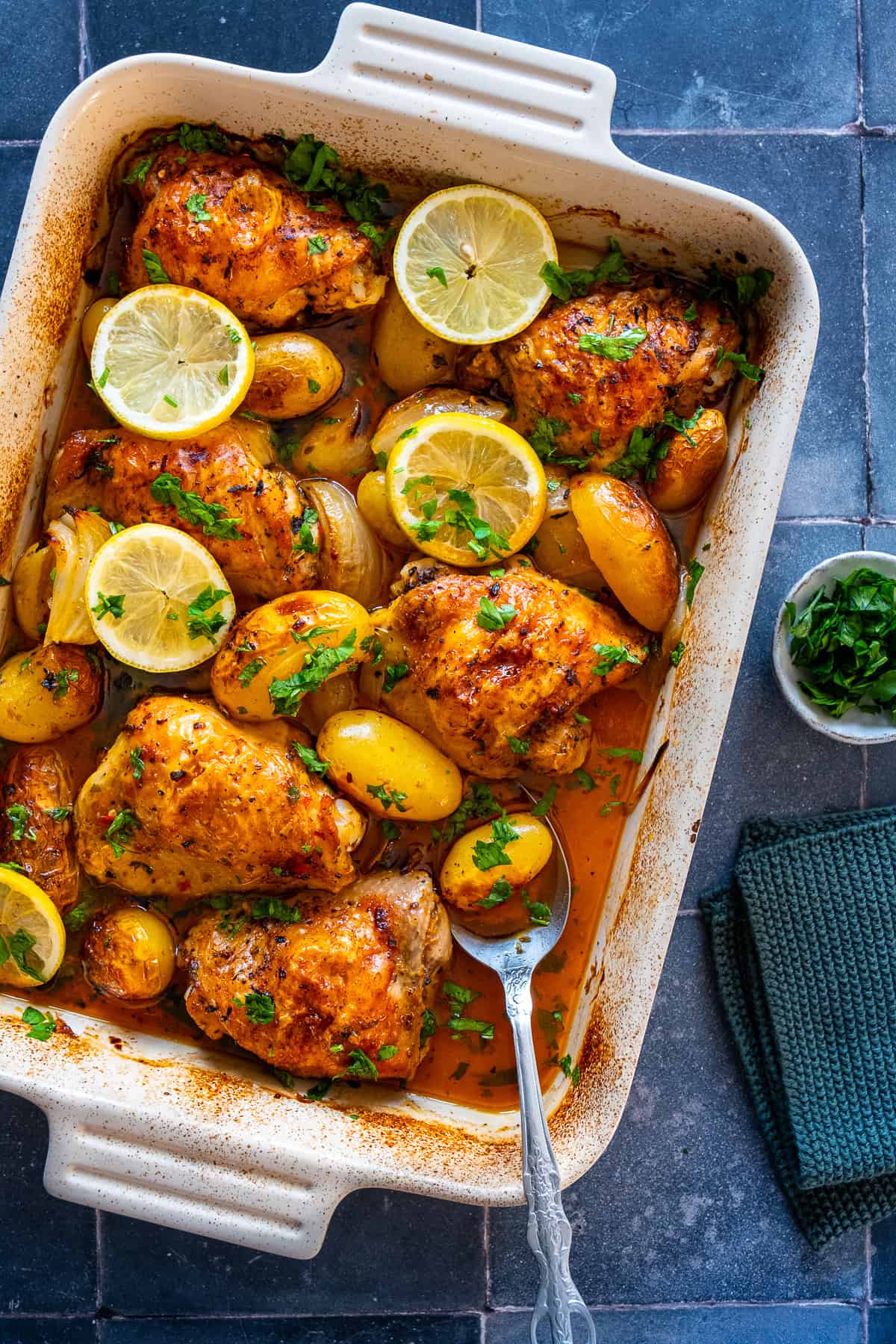 Overhead view of baked lemon garlic chicken thighs and baby potatoes in a white dish, garnished with lemon slices and parsley, with a spoon and napkin on the side.