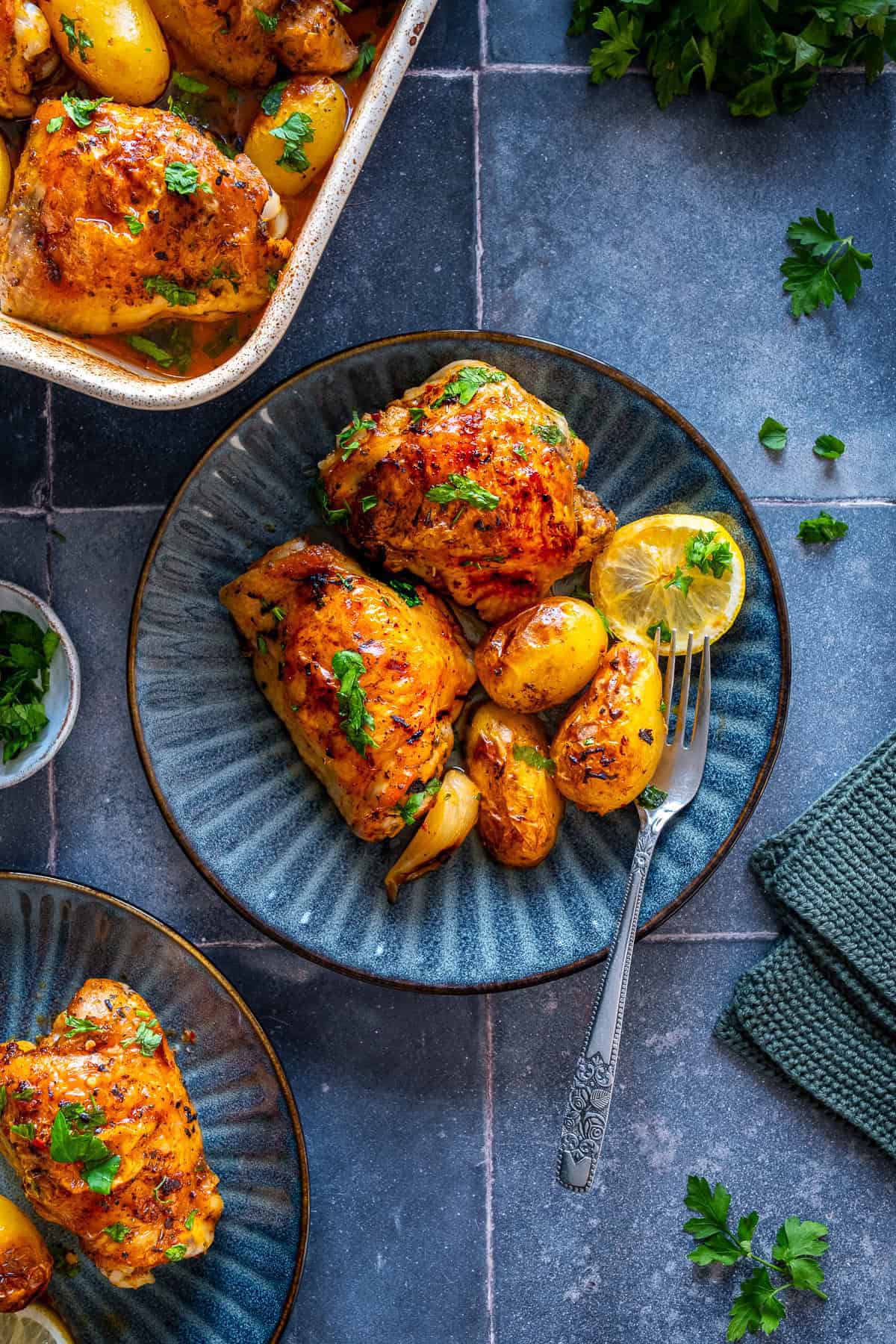 Plated serving of lemon garlic chicken thighs and baby potatoes on a blue plate with lemon slice and parsley, next to the baking dish.