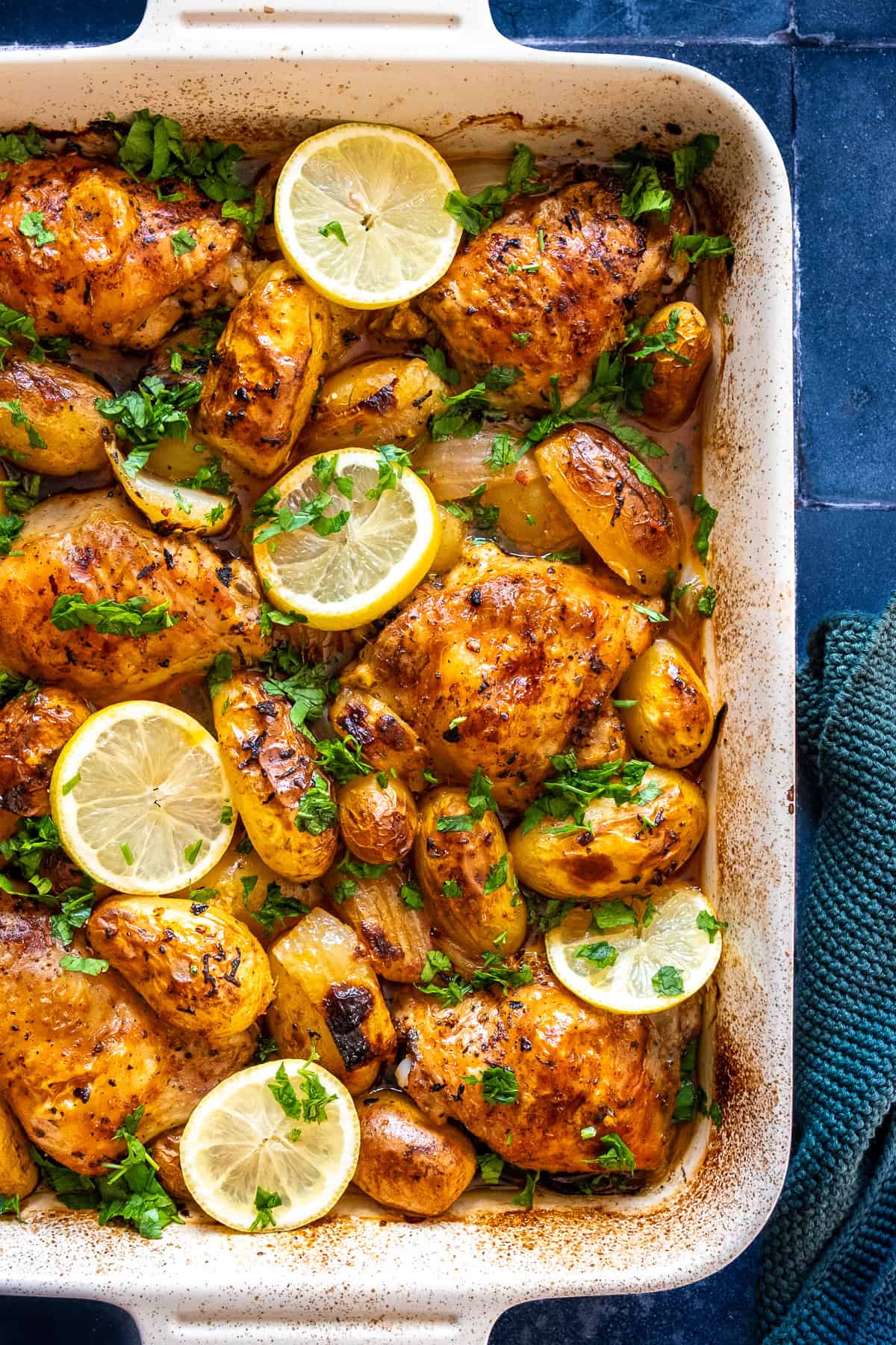 Baked lemon garlic chicken thighs and baby potatoes in a white dish, topped with lemon slices and fresh parsley.