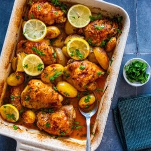Baked lemon garlic chicken thighs with baby potatoes in a white baking dish, garnished with lemon slices and fresh parsley.