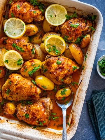 Baked lemon garlic chicken thighs with baby potatoes in a white baking dish, garnished with lemon slices and fresh parsley.
