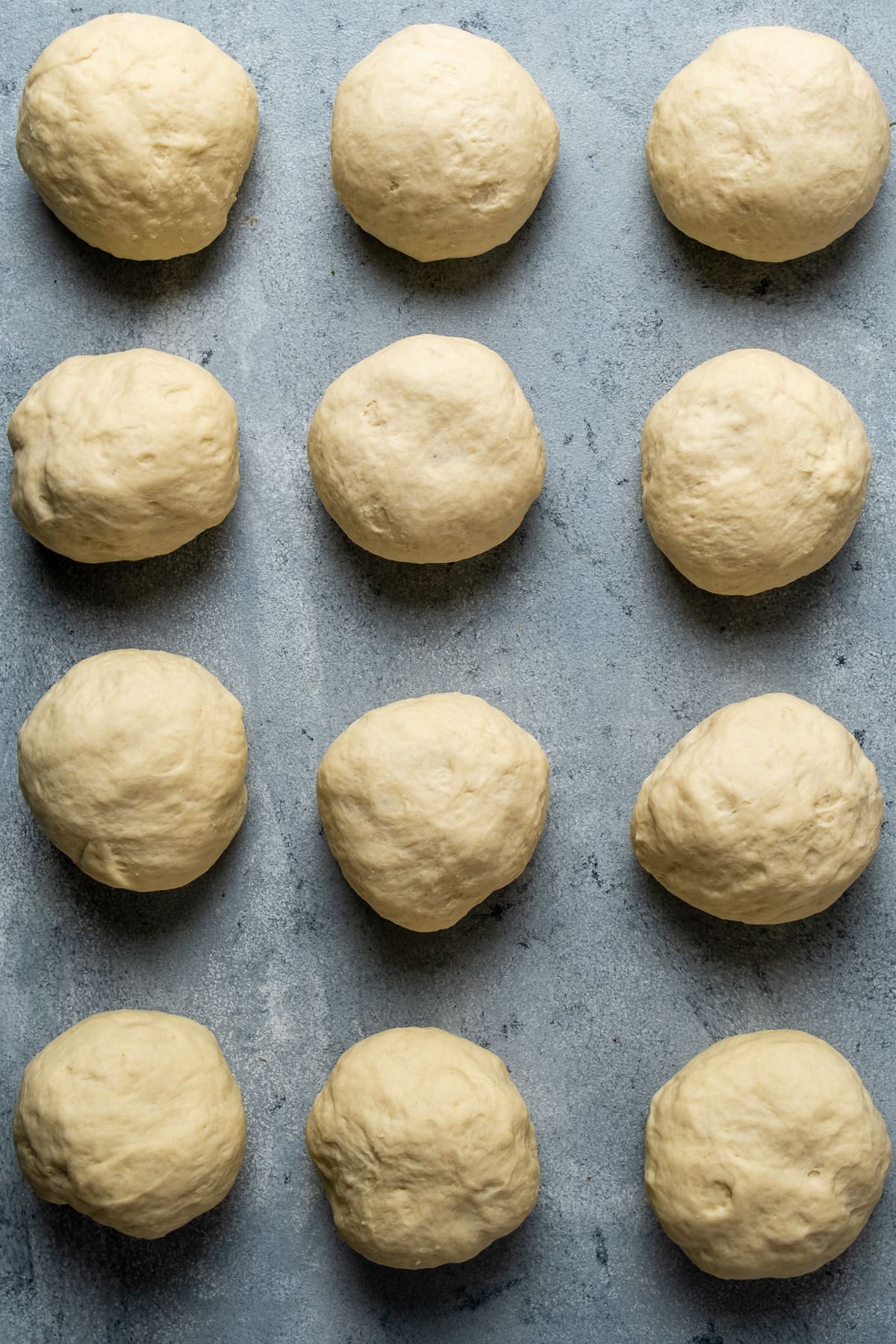 Twelve mini dough balls are aligned in four rows and three coloumns on a grey background.